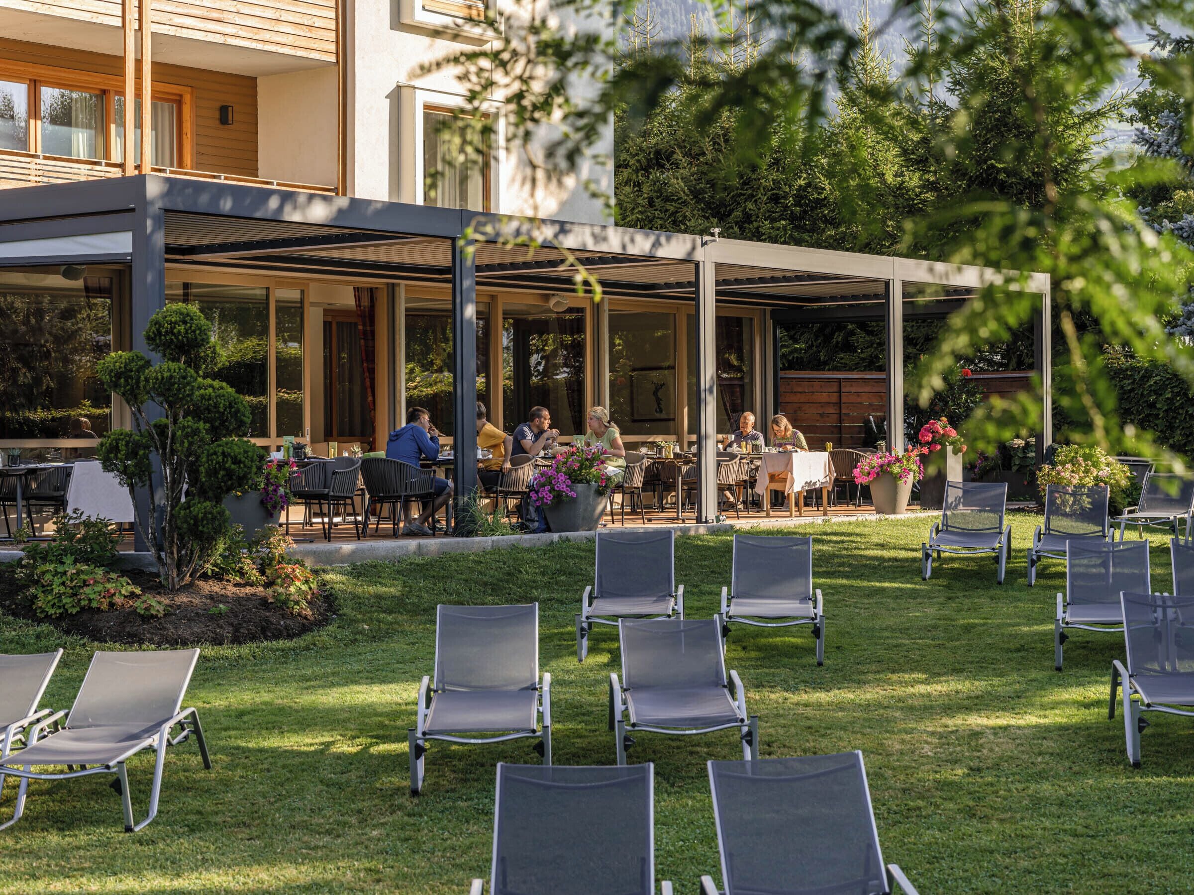 An outdoor patio area with tables and chairs, surrounded by greenery and a building with large windows.