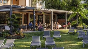 An outdoor patio area with tables and chairs, surrounded by greenery and a building with large windows.