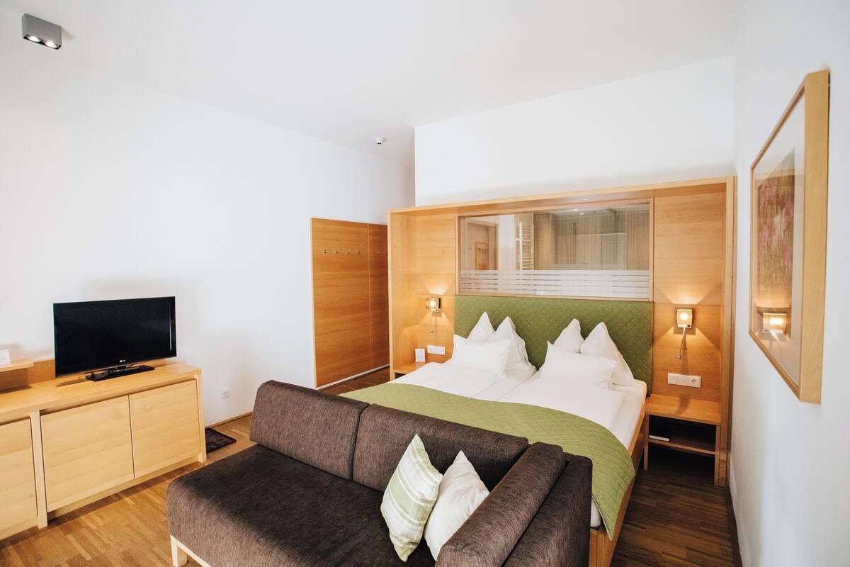 Ein modernes Hotelzimmer mit einem Bett, einem Fernseher auf einer Holzkommode, einem grünen Sofa und zwei Nachttischlampen.