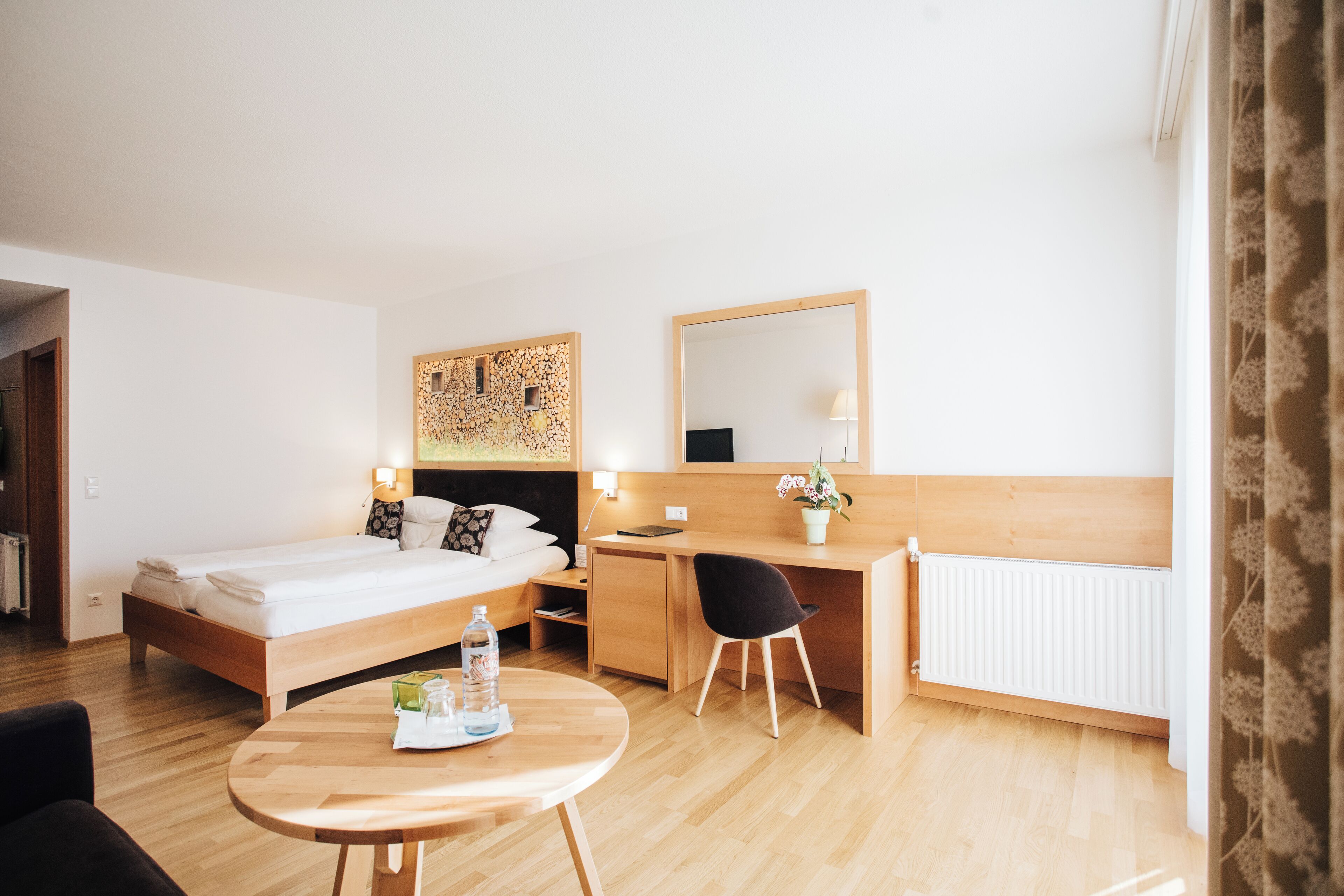 A hotel room with a bed, desk, chair, and a small table with a bottle and glasses.
