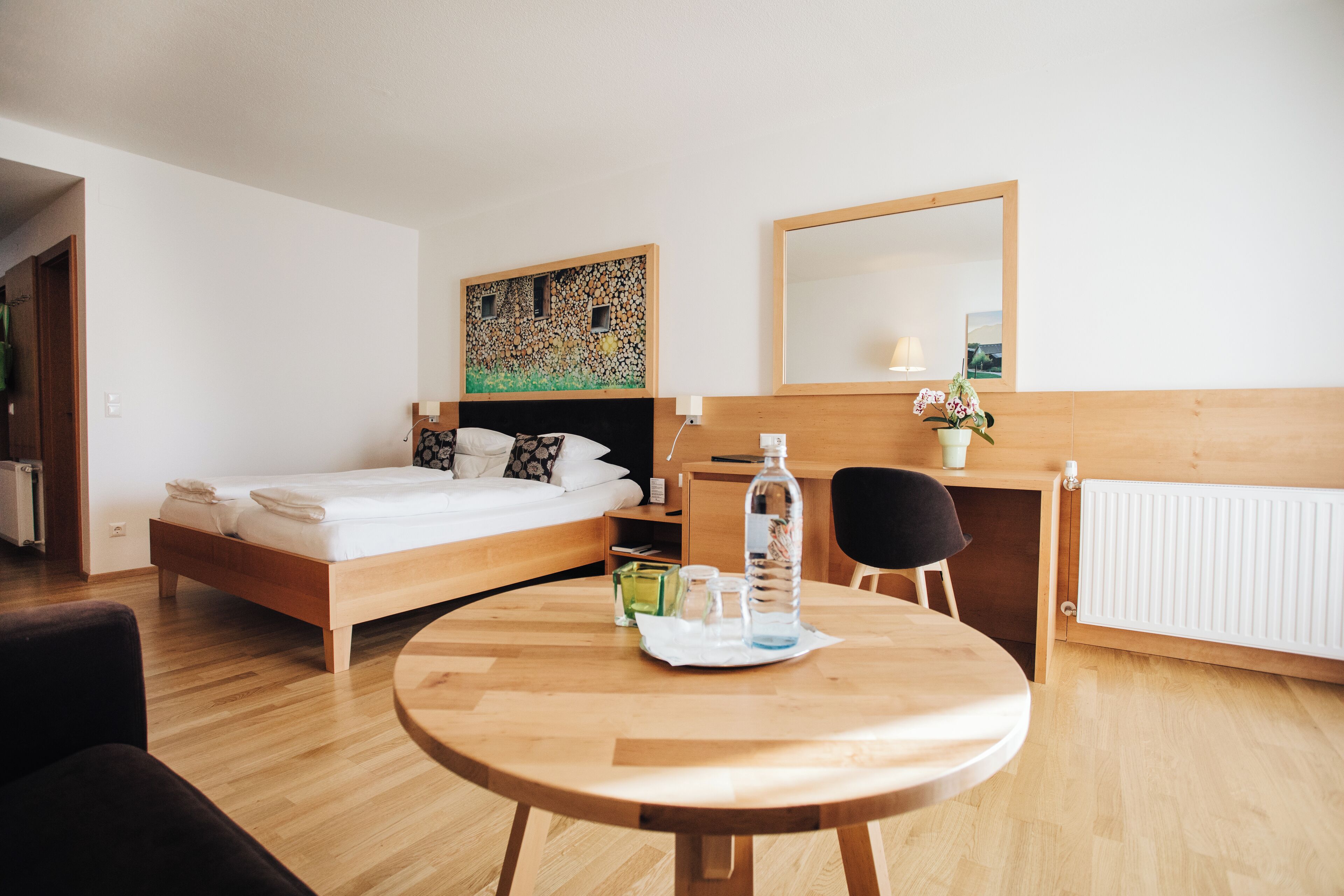 A hotel room with a bed, a desk, a chair, a round table with a bottle and glasses, a painting on the wall, and a radiator.