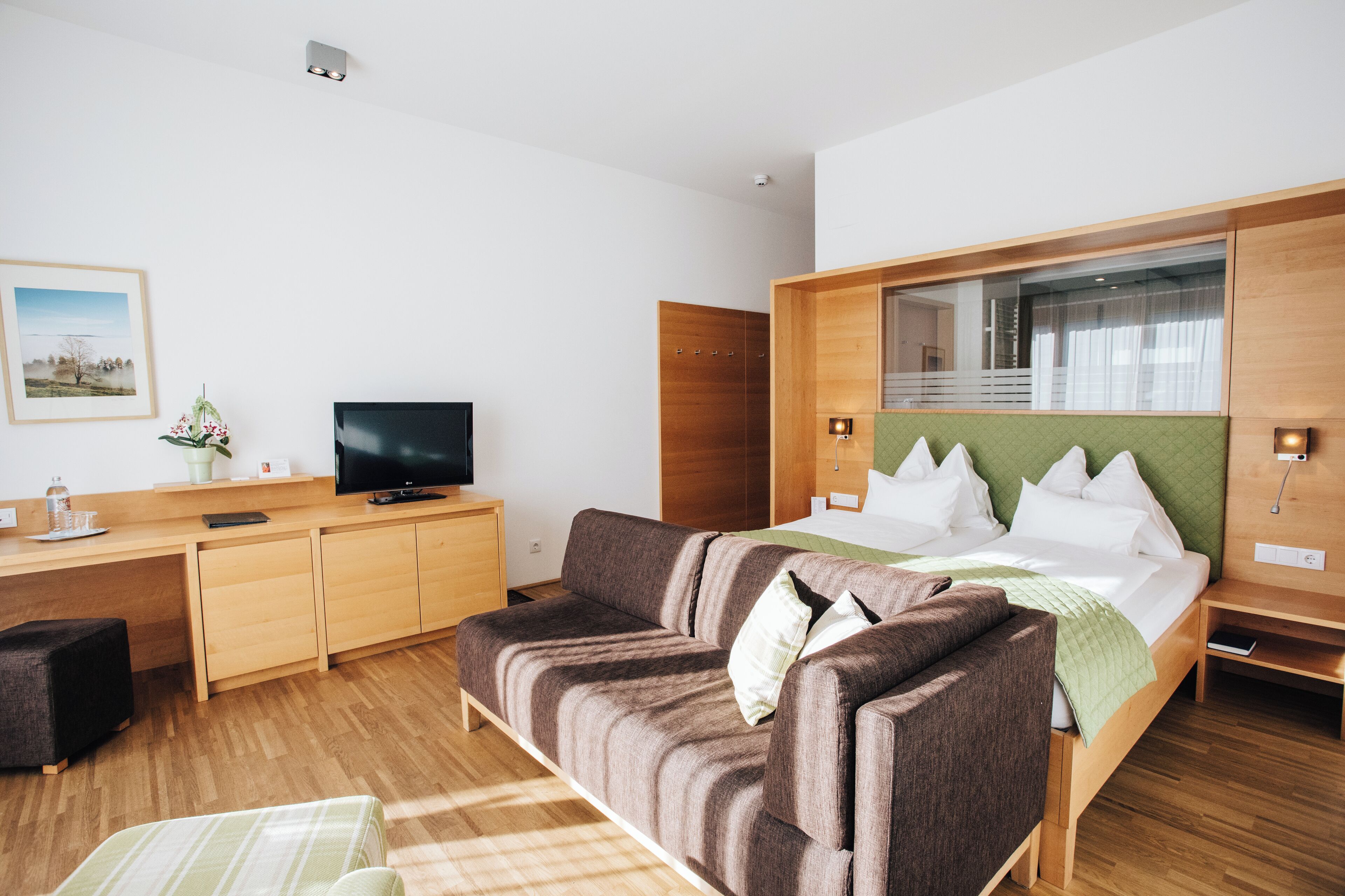 A modern hotel room with a wooden desk, a flat-screen TV, a sofa, and a bed with a green headboard.
