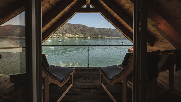 A wooden deck with two lounge chairs facing a lake with boats and a hilly landscape in the background.