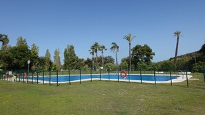 A swimming pool surrounded by a green fence, with palm trees and a clear blue sky in the background. - Luxury Apartment San Roque Club. Spacious, large balcony overlooking mountains (San Roque)