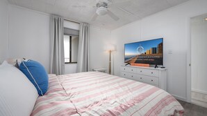 A hotel room with a bed, a television displaying a cityscape, a window with curtains, and a ceiling fan. - New For 2025! Charming 1 bedroom condo oceanblock in North OC (Ocean City)