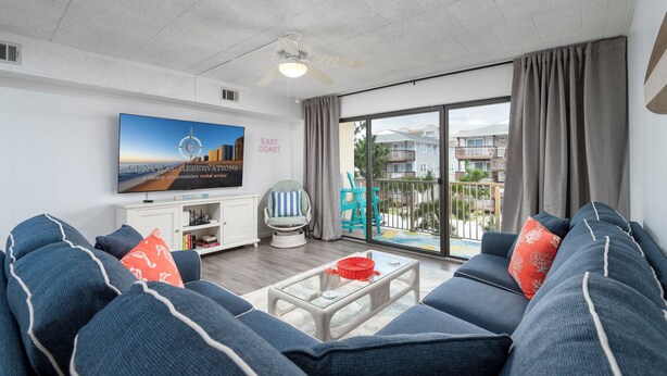 A modern living room with a large flat-screen TV, a comfortable sofa set, a glass coffee table, and a view of the outside through a sliding glass door. - New For 2025! Charming 1 bedroom condo oceanblock in North OC (Ocean City)