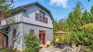 A two-story house with a red door and windows, a patio with a table and chairs, and a garden with various plants and a tree. - Cliffwood Cottage by Evergreen Home (Lenox)