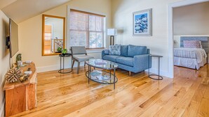 A spacious living room with hardwood flooring, a grey sofa, a round coffee table, and a large window with blinds. - Cliffwood Cottage by Evergreen Home (Lenox)