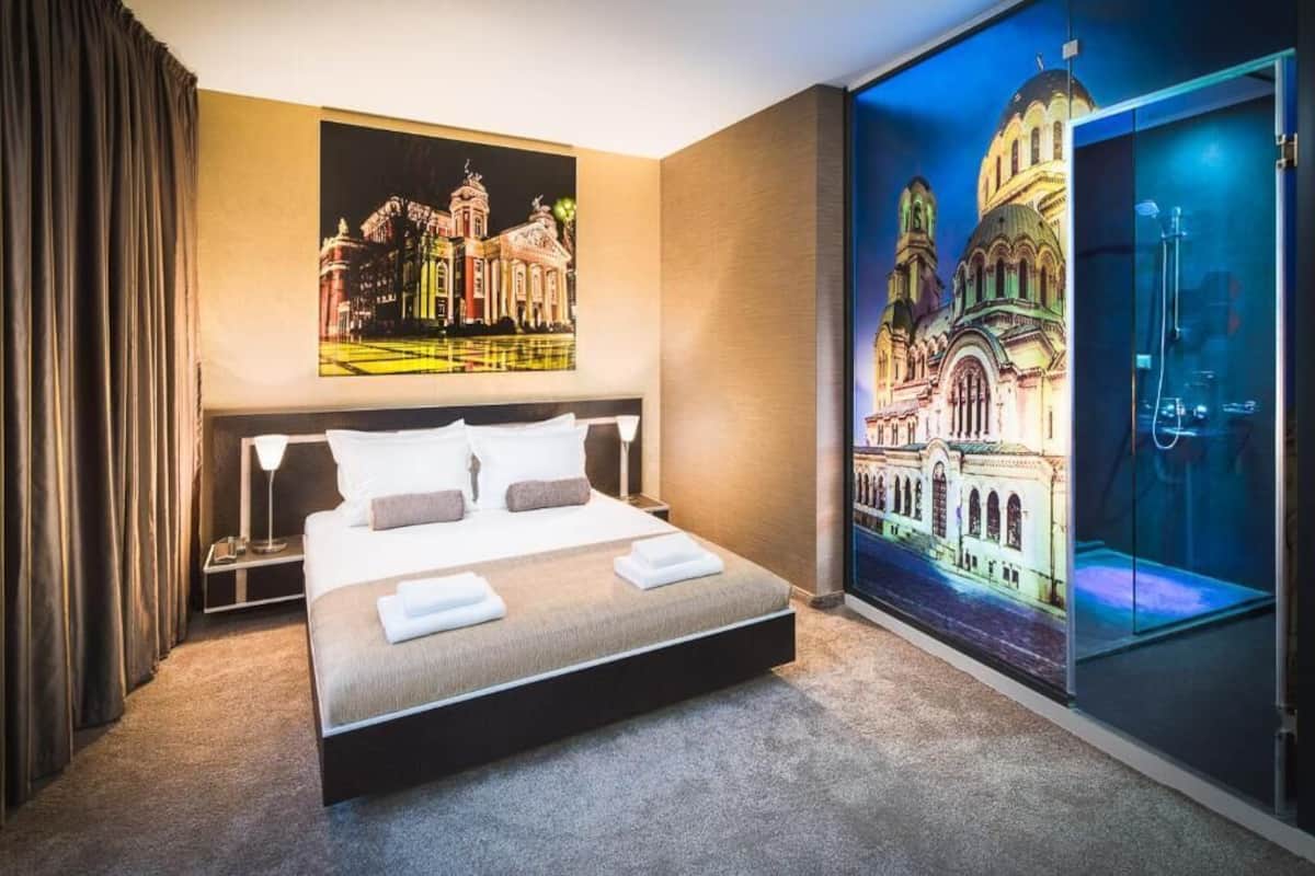 A hotel room with a large bed, a nightstand, a wall-mounted TV displaying a cityscape, and a glass door leading to a bathroom.