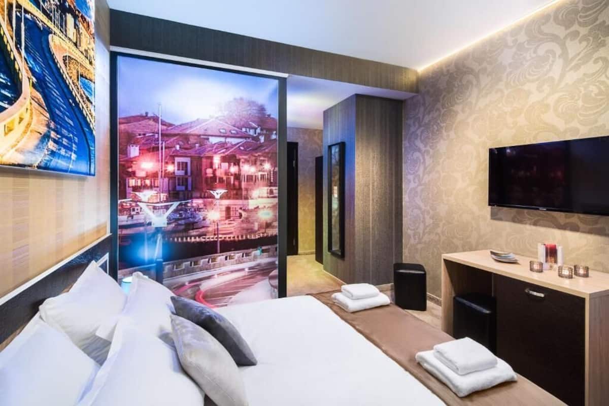 A modern hotel room with a large bed, a TV, a desk, and a view of a cityscape.