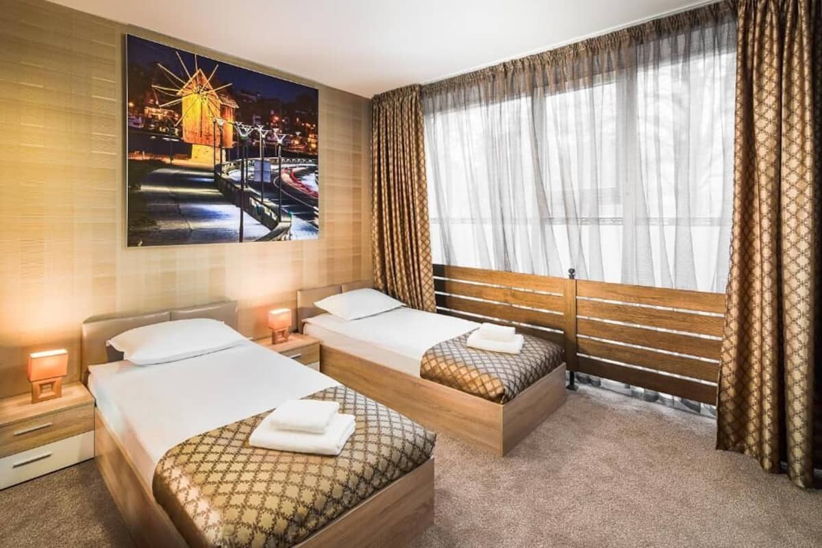 A hotel room with two beds, a large window with sheer curtains, a cityscape painting on the wall, and a night view of a bridge.