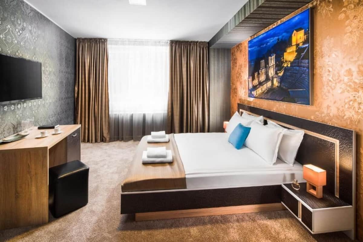 A hotel room with a large bed, a desk with a flat-screen TV, a painting on the wall, and a window with curtains.