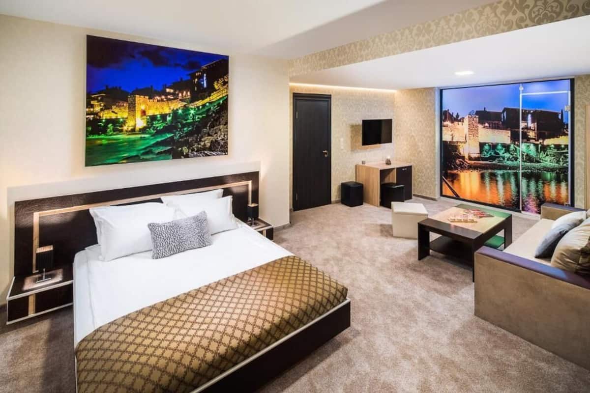 A hotel room with a large bed, a sofa, a small table, a TV, and a large window with a cityscape view.