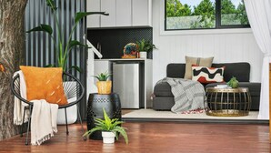 Una sala de estar moderna con un sillón negro, un sofá y una mesita. Se ve una ventana grande con vistas a árboles y una cortina blanca.
