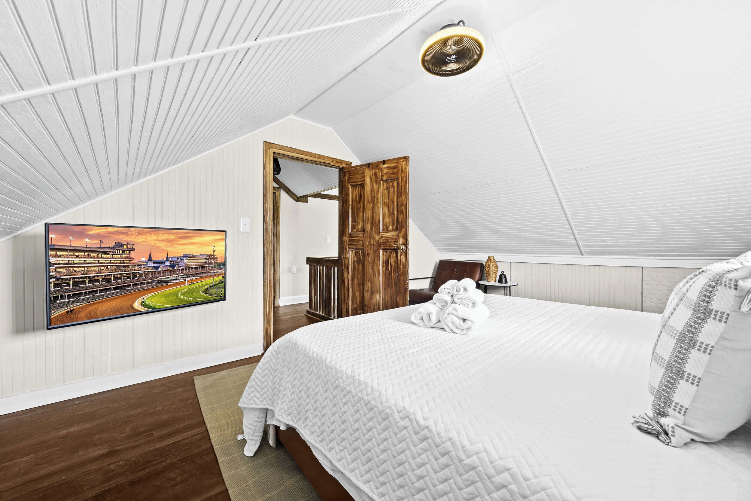 Une chambre à coucher avec un grand lit, une porte en bois, un tableau représentant une piste de course et un ventilateur de plafond.