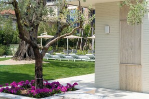 A tree-lined outdoor seating area with lounge chairs and umbrellas, a grassy lawn, and a building with a wooden door.