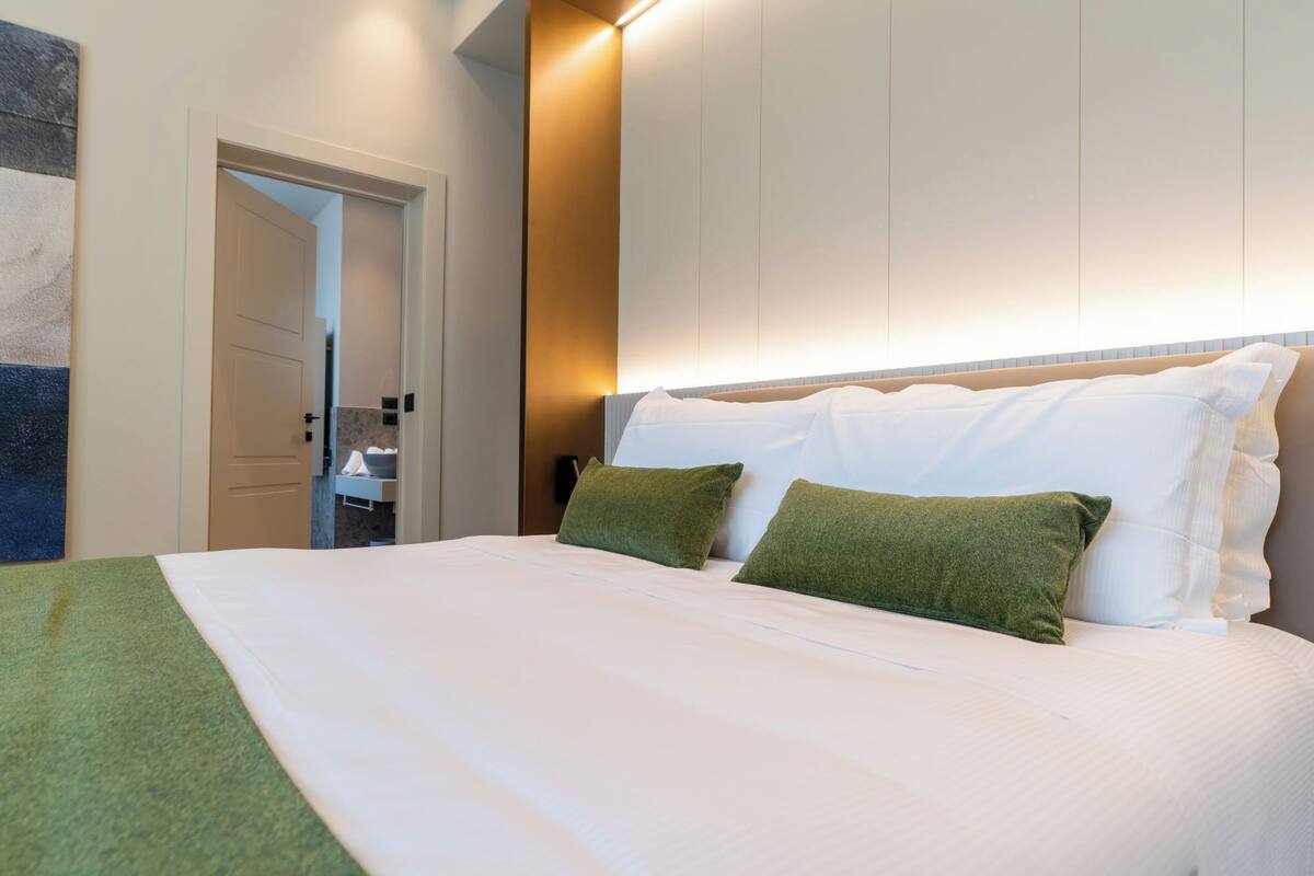 A neatly made bed with white and green pillows, a headboard with built-in lighting, and a door leading to a bathroom.
