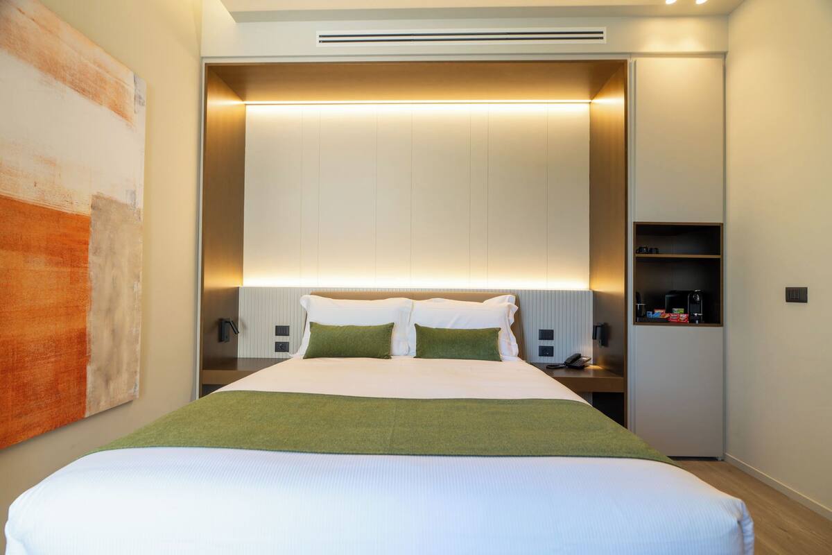 A modern hotel room with a large bed, a bedside table with a phone, and a shelf with toiletries.
