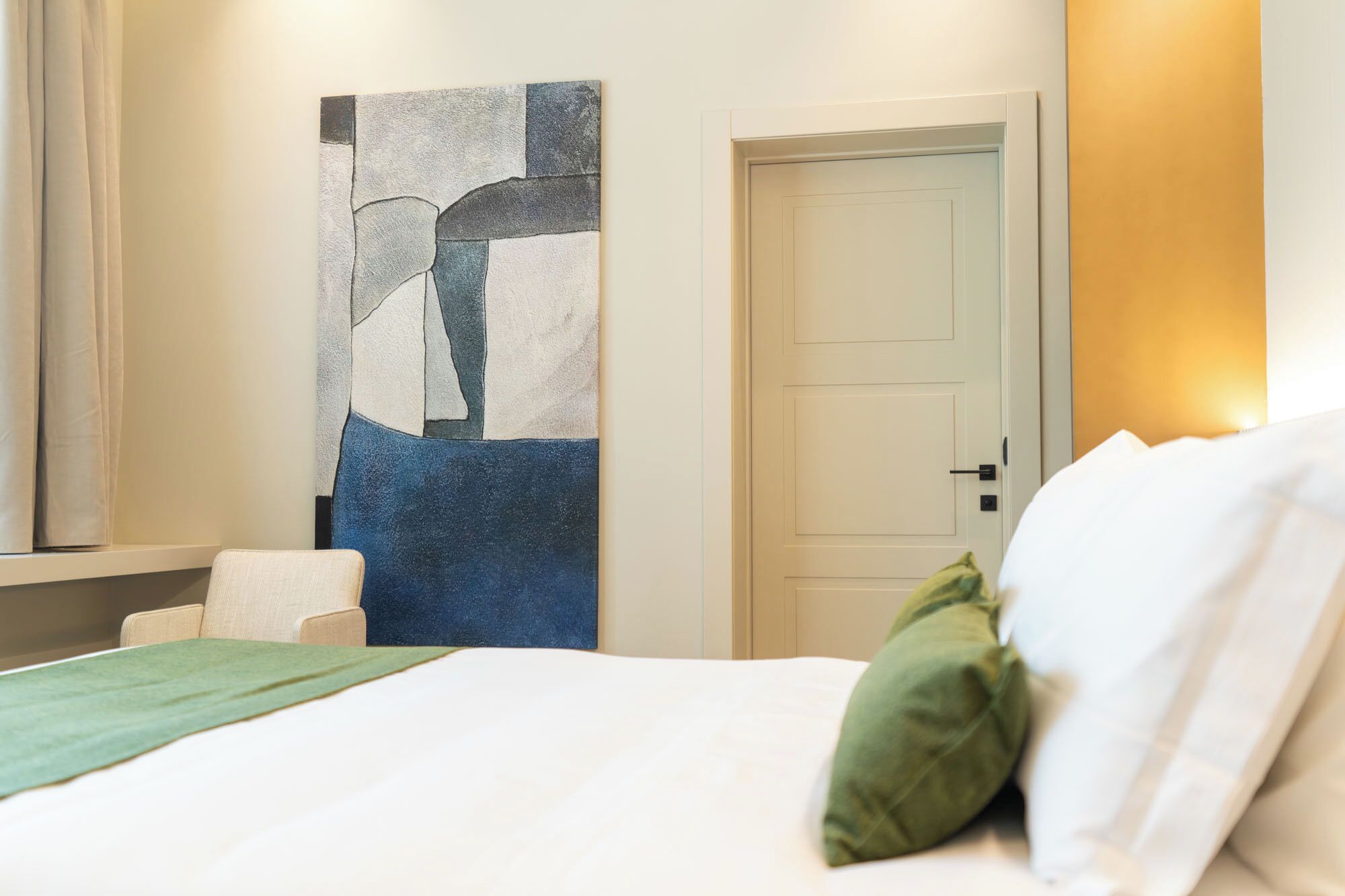 A hotel room with a bed, a green throw pillow, a white pillow, a painting on the wall, and a door.