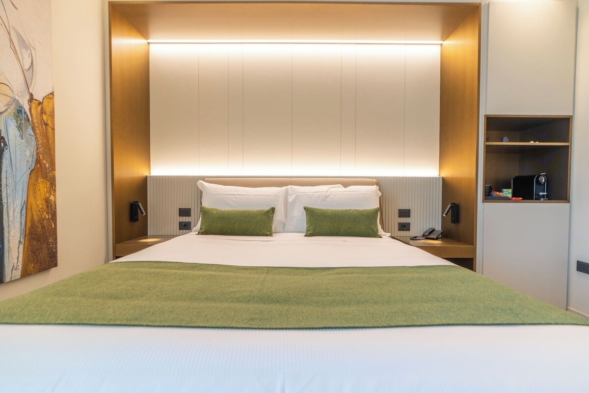 A modern hotel room with a large bed, two green throw pillows, a bedside table with a lamp, and a wall-mounted shelf with a coffee machine.