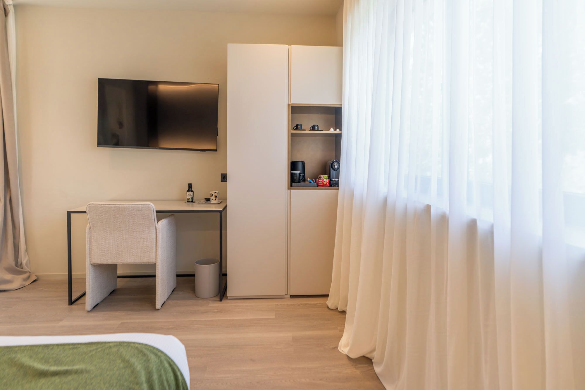 A hotel room with a bed, a desk with a chair, a TV mounted on the wall, a minibar, and sheer curtains.