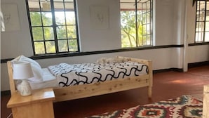 A room with a wooden bed, a bedside table with a lamp, a patterned rug, and windows with views of greenery. - Uncle Bob's Farm Hotel Nyeri/Nanyuki (Nairobi)