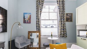 A living room with a grey armchair, a round table with a bottle and glasses, a mirror, and a sofa with yellow cushions. - Chic flat in Central London near Victoria Station - SW1 (London)