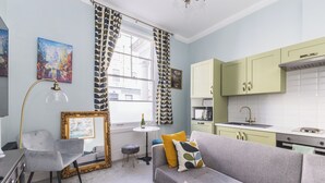 A compact living space with a grey sofa, a small round table, and a chair. There is a window with patterned curtains, a painting on the wall, and a kitchen area with green cabinets. - Chic flat in Central London near Victoria Station - SW1 (London)