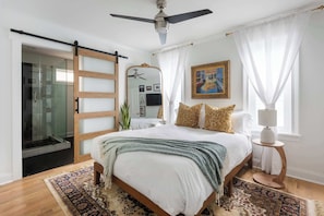 A bedroom with a bed, a ceiling fan, a large mirror, a painting, and a window with curtains.
