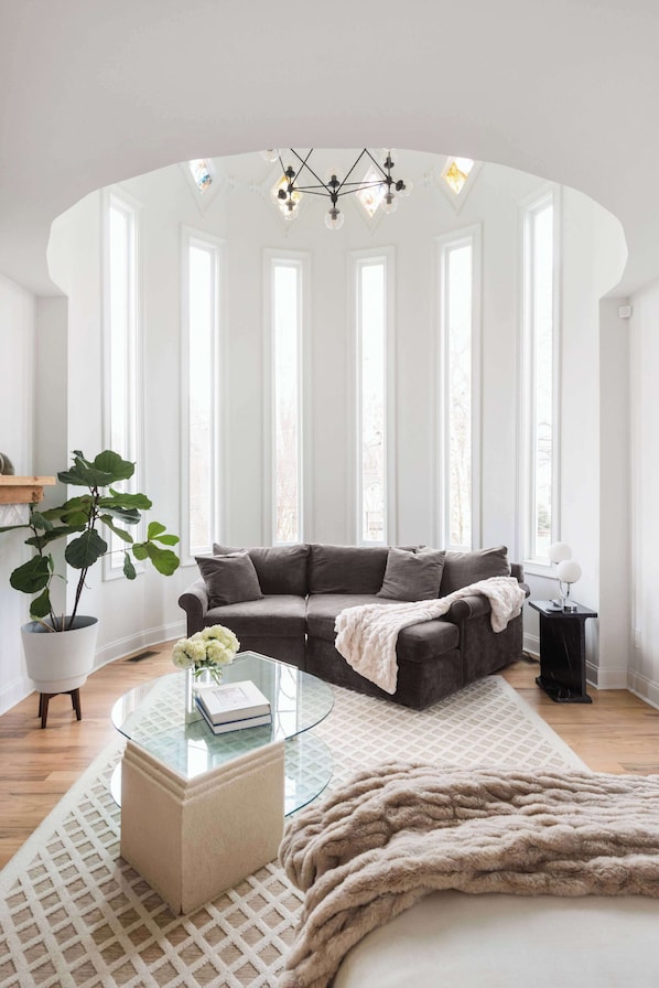 A modern living room with a dark grey sofa, a glass coffee table, a large window with multiple panes, and a hanging light fixture.