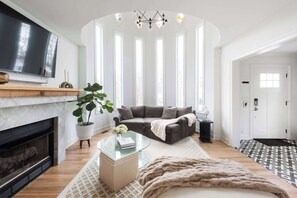Ein geräumiges Wohnzimmer mit Kamin, einem großen Flachbildfernseher, einem grauen Sofa, einem Glastisch und einem großen Fenster mit vertikalen Jalousien.