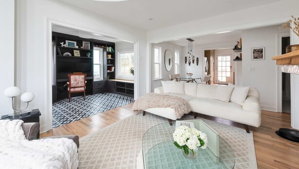 Ein geräumiges Wohnzimmer mit einem weißen Sofa, einem Glastisch mit Blumen, einem gemusterten Teppich und einem schwarz-weißen Fußboden.