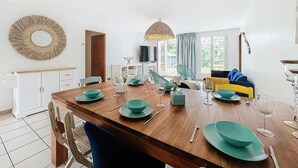 Ein Essbereich mit einem Holztisch, gedeckt für vier Personen, umgeben von Stühlen. Ein Fernseher ist an der Wand montiert, und es gibt ein Fenster mit Vorhängen.