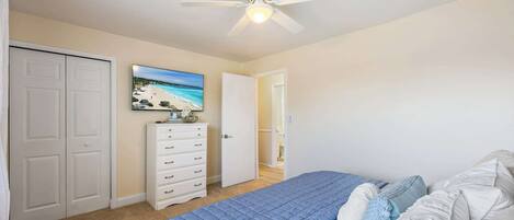 Un dormitorio con cama, ventilador de techo, cómoda, armario y un cuadro de una playa.