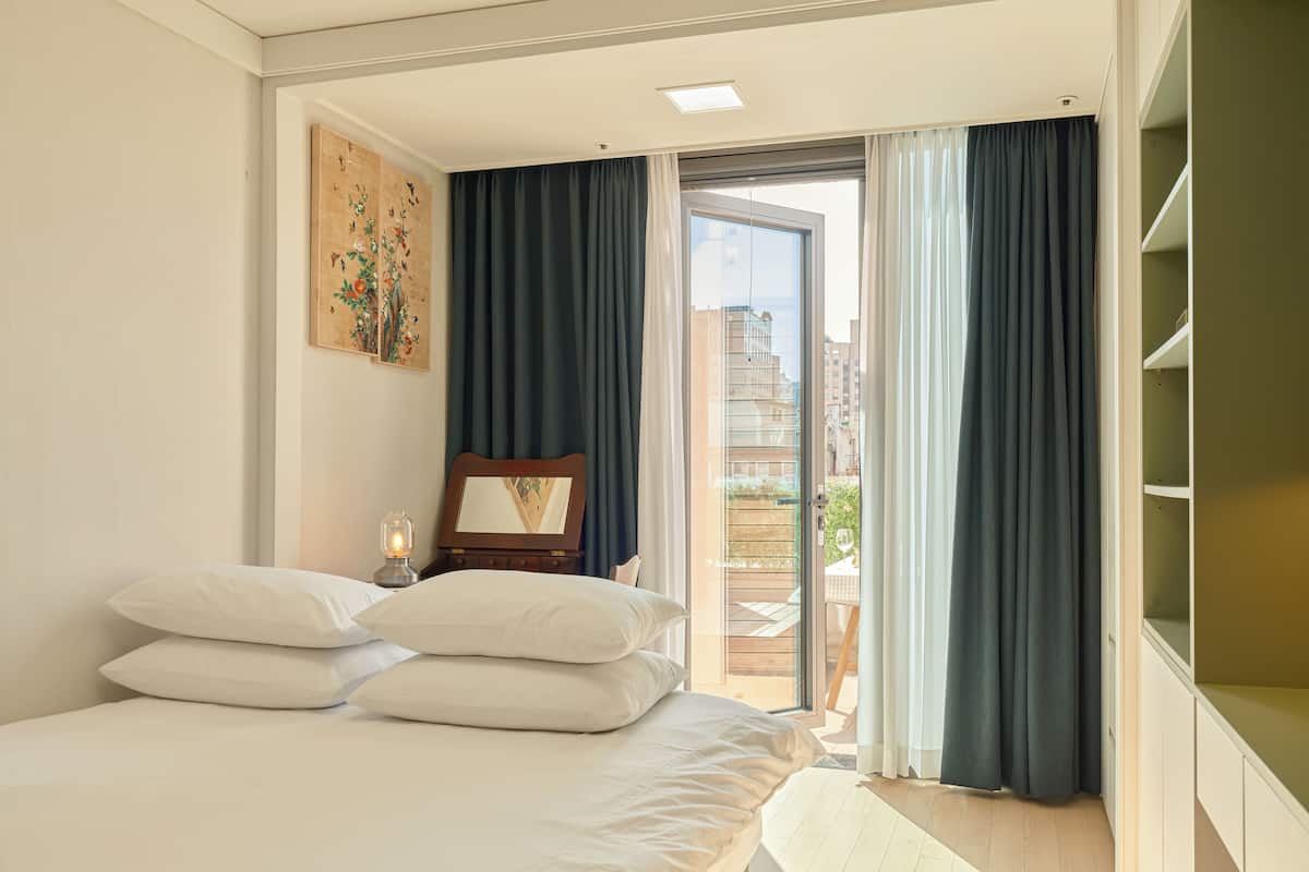 A bedroom with a bed, a nightstand with a lamp, a mirror, and a view of the outside through a sliding glass door.
