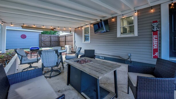 An outdoor patio with a wooden table, wicker chairs, and a TV mounted on the wall. - Tide on 19th- 4BR/2BA, Large Patio, Fire Pit & Smart TVs! (Northport)