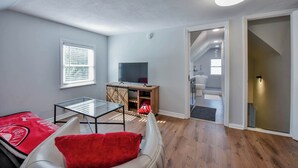 A living room with a glass coffee table, a red throw pillow, a television on a wooden stand, and a view into the kitchen. - Tide on 19th- 4BR/2BA, Large Patio, Fire Pit & Smart TVs! (Northport)