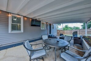 A covered patio area with a round table and chairs, a grill, and string lights. - Tide on 19th- 4BR/2BA, Large Patio, Fire Pit & Smart TVs! (Northport)