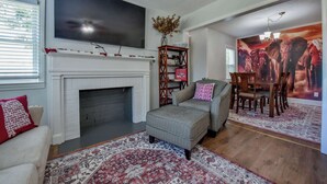 A living room with a fireplace, a television, a bookshelf, and a dining area with a large elephant mural. - Tide on 19th- 4BR/2BA, Large Patio, Fire Pit & Smart TVs! (Northport)