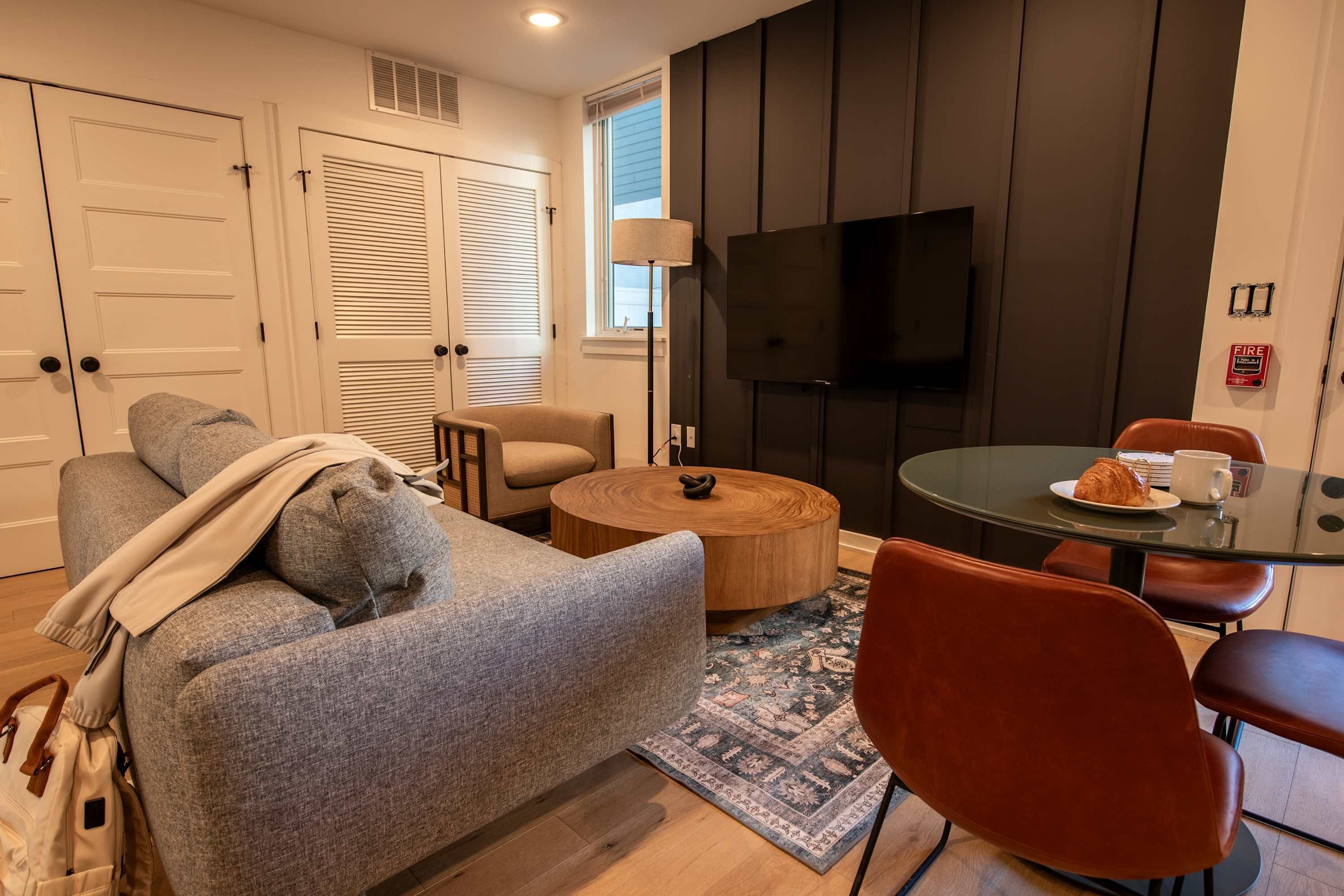 A modern living room with a grey sofa, a round wooden coffee table, and a red chair. There is a television mounted on the wall, a small table with a plate and a cup, and a patterned rug on the floor.