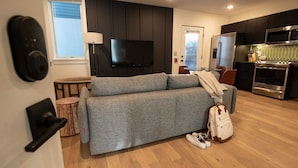 A modern living room with a grey sofa, a flat-screen TV, a wooden floor, and a kitchen area with a microwave and oven.