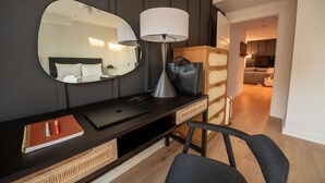 A modern hotel room with a dark wood desk, a large mirror, a lamp, a laptop, a notebook, and a chair.