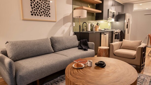 Ein modernes Wohnzimmer mit einem grauen Sofa, einem runden Couchtisch aus Holz und einem beigen Sessel.
