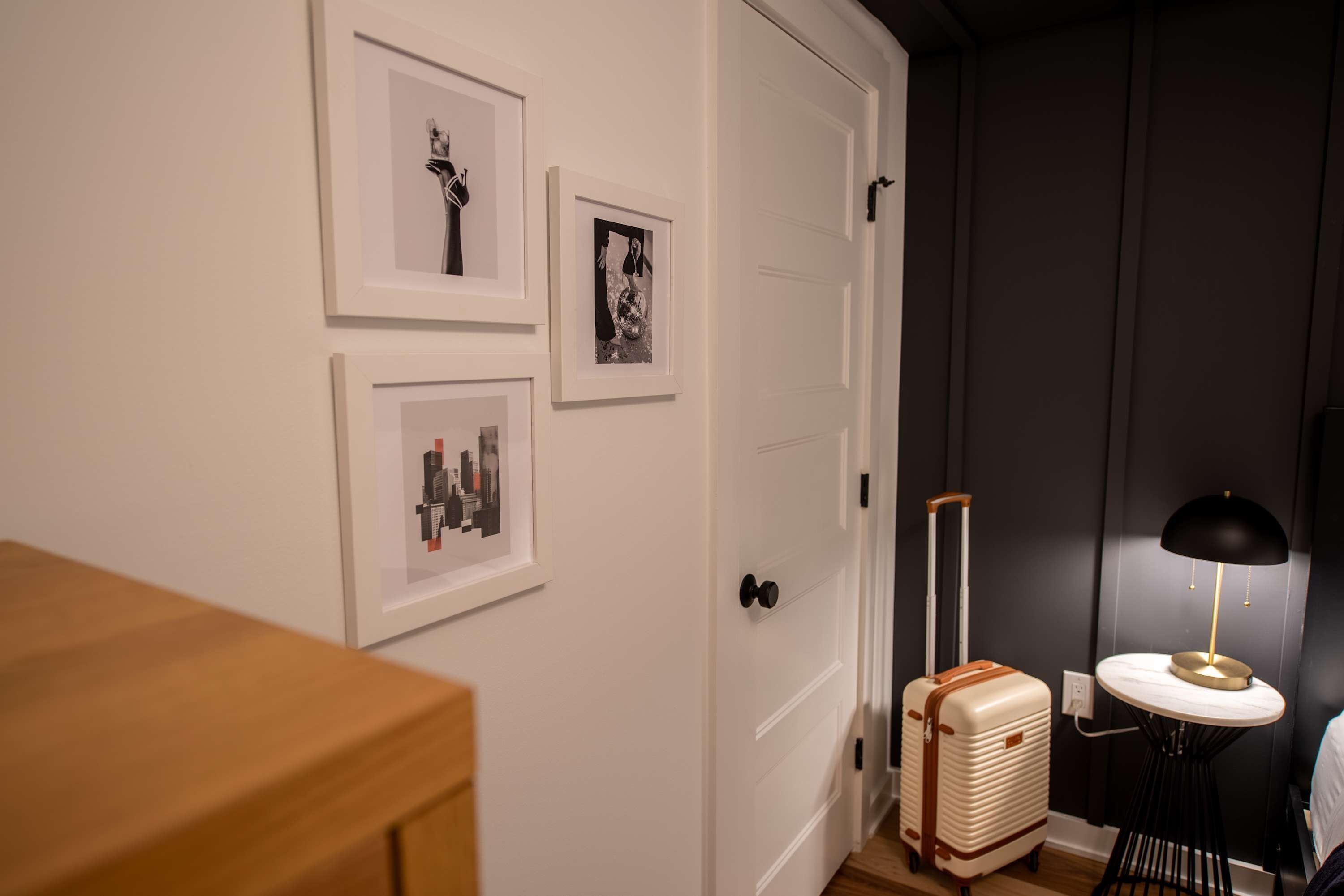 A room with a wooden dresser, a white door, and a wall with framed pictures. There is a vintage suitcase and a small table with a lamp.