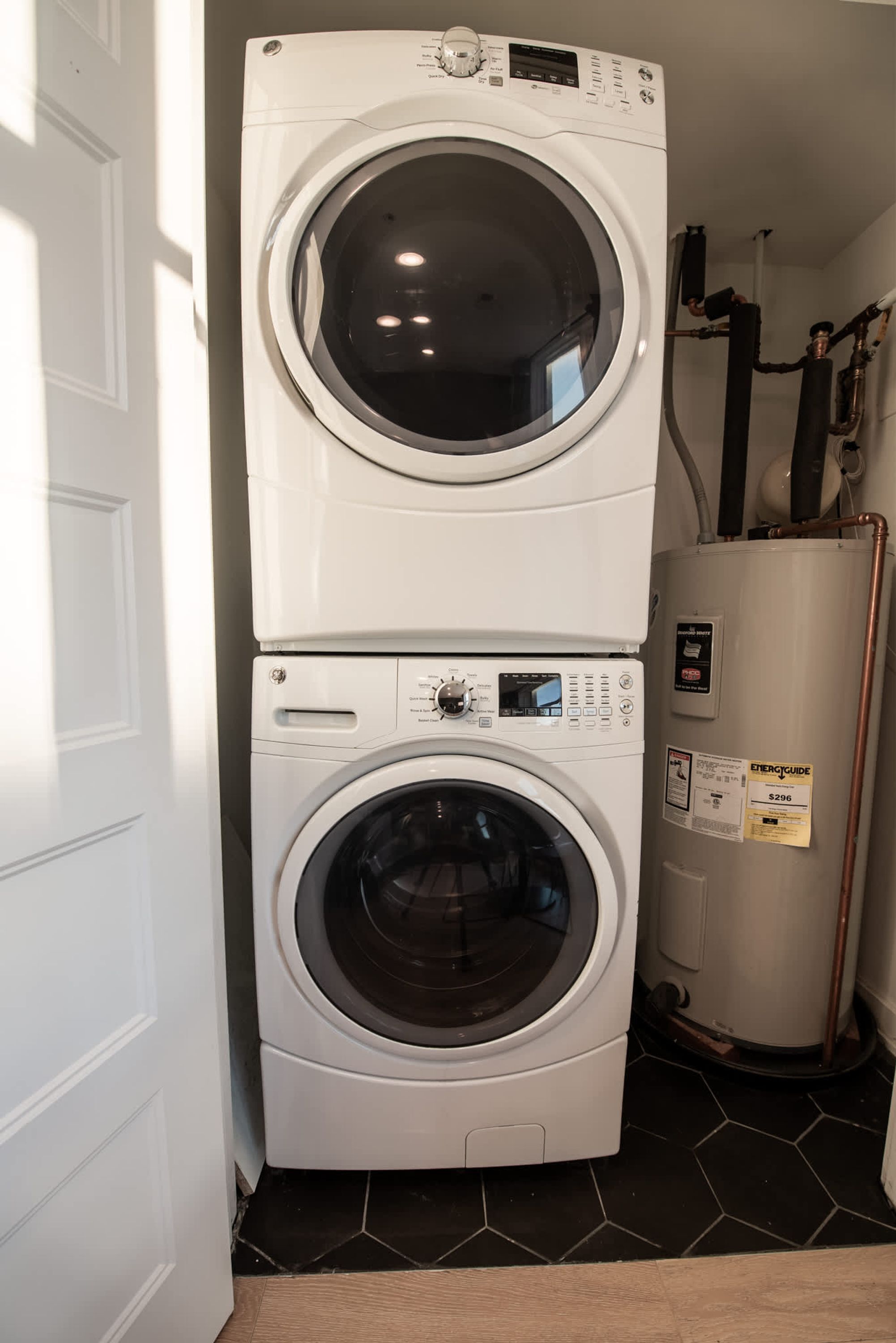 A stacked washer and dryer unit in a laundry room with a tiled floor and a water heater nearby.