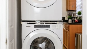 Un appareil combiné lave-linge et sèche-linge, installé en hauteur, dans une cuisine dotée de meubles en bois et d'une poubelle en acier inoxydable.