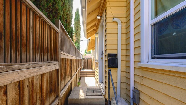 Un étroit sentier en bois mène à une maison jaune, avec une boîte aux lettres et une fenêtre ornée de vitraux.