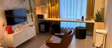 Una habitación de hotel moderna con televisión de pantalla plana, microondas, refrigerador pequeño, sillón de cuero, alfombra redonda y mesita de noche con lámpara.
