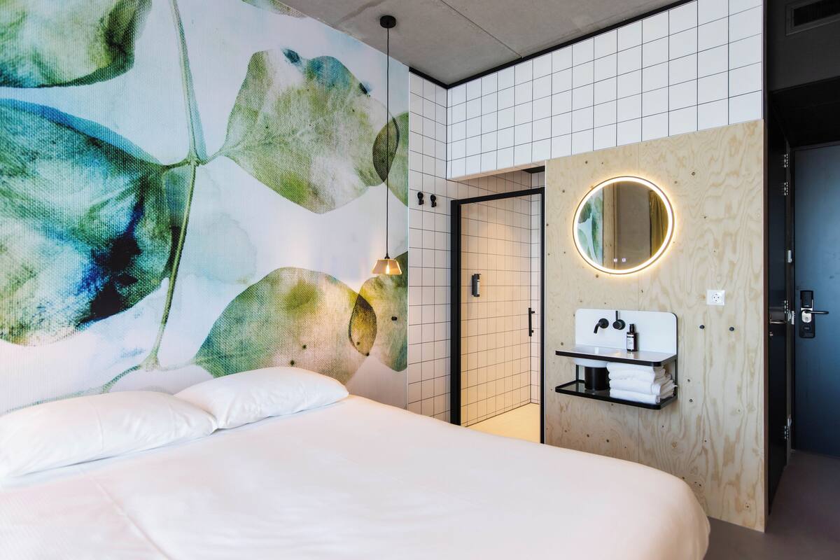 Une chambre d’hôtel moderne dotée d’un grand lit, d’un grand miroir rond fixé au mur et d’un panneau en bois intégrant un lavabo.