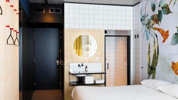 A modern hotel room with a wooden wardrobe, a round mirror above the sink, a bed with white linens, and a floral mural on the wall.